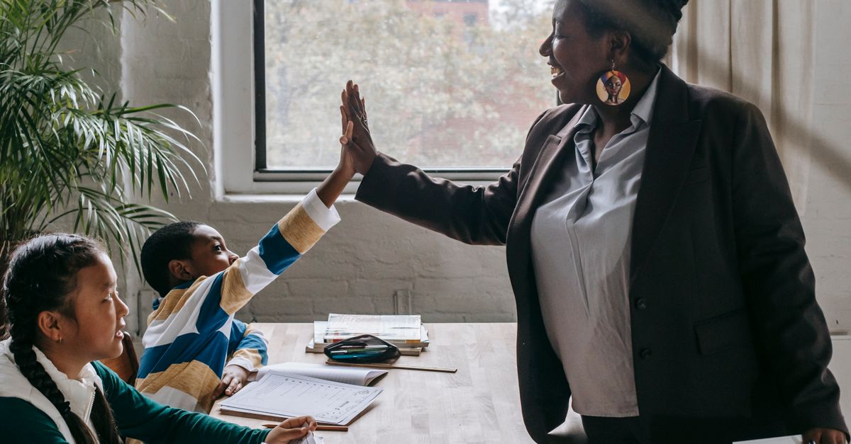 Side view of smiling black boy giving high five to teacher while sitting with diverse classmate girl at desk in classroom